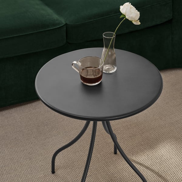 Table d’appoint ronde noire avec 3 pieds, boisson et fleur, sur tapis beige, à côté d’un canapé vert.