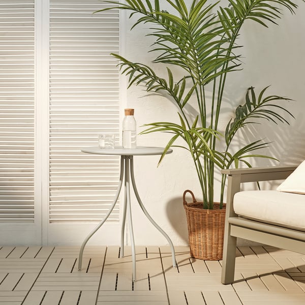 La petite table d’appoint beige TÅNEBRO avec plateau blanc et pieds minces se trouve près d’une grande plante dans un panier tressé et une chaise sur un patio extérieur en bois.