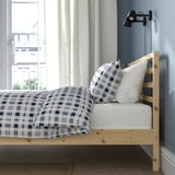 Un cadre de lit en bois soigneusement conçu avec une couette à motifs à carreaux et des oreillers contre une fenêtre, avec un support à lattes et un design épuré.