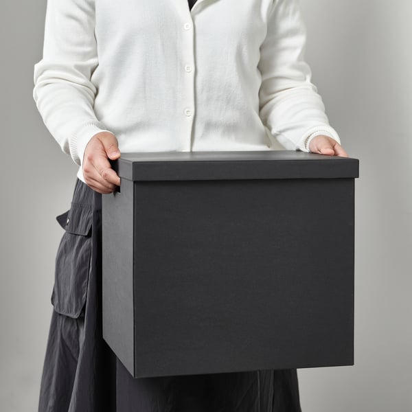 Personne tenant une grande boîte TJENA grise rectangulaire ; Idéal pour le stockage de gadgets multimédias ou d’articles de loisirs.