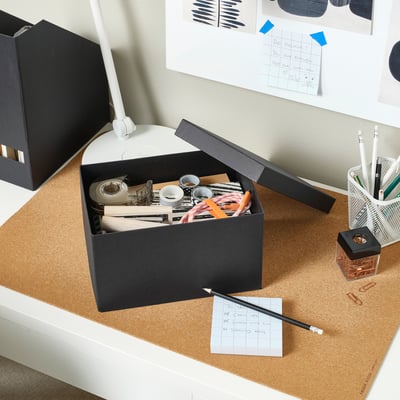 Configuration soignée du bureau avec boîte noire TJENA pour ranger les fournitures, près d’un moniteur et d’un tableau en liège.