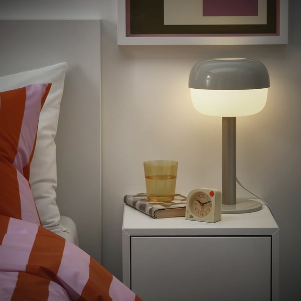 Chambre confortable avec une table de chevet moderne, une lampe grise, une horloge jaune, un verre d’eau et un livre plié, créant une atmosphère calme.