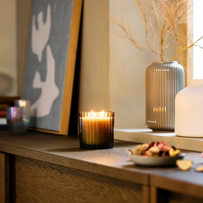 Un espace de vie douillet avec une bougie parfumée à TOFSTÄRNA . Bougie allumée sur l’étagère avec décor, vases et art abstrait.