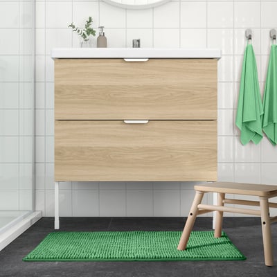 Salle de bain moderne avec vanité en bois clair, comptoir et lavabo blancs. Le tapis TOFTBO vert, les serviettes et le banc sont soigneusement disposés. Le tapis est petit.
