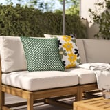 Chaise rembourrée blanche avec des coussins en losange vert et des fleurs jaunes, entourée de verdure.