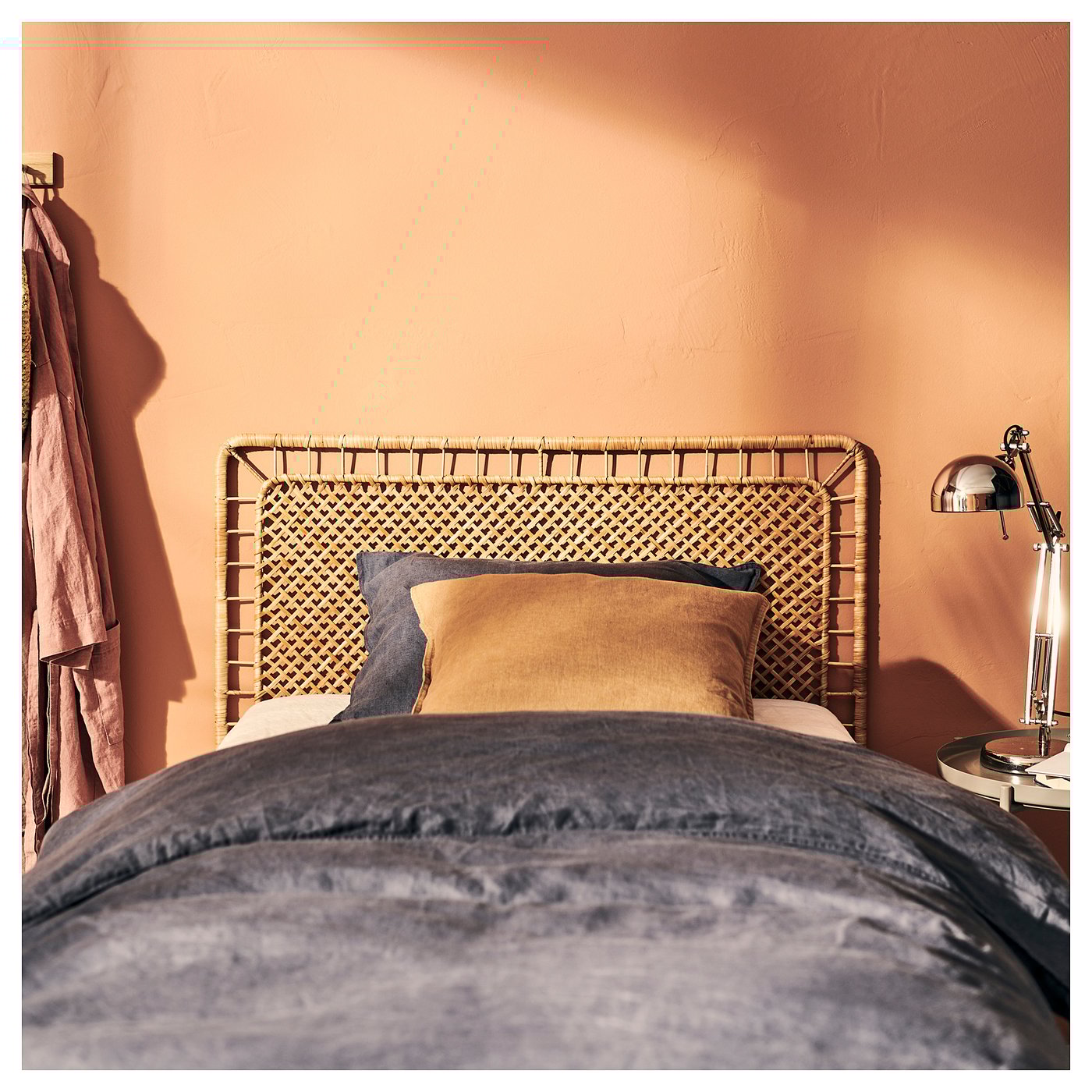 Tête de lit en rotin naturel avec motif tissé, oreillers beiges, mur pêche, literie grise, lampe, cintre.