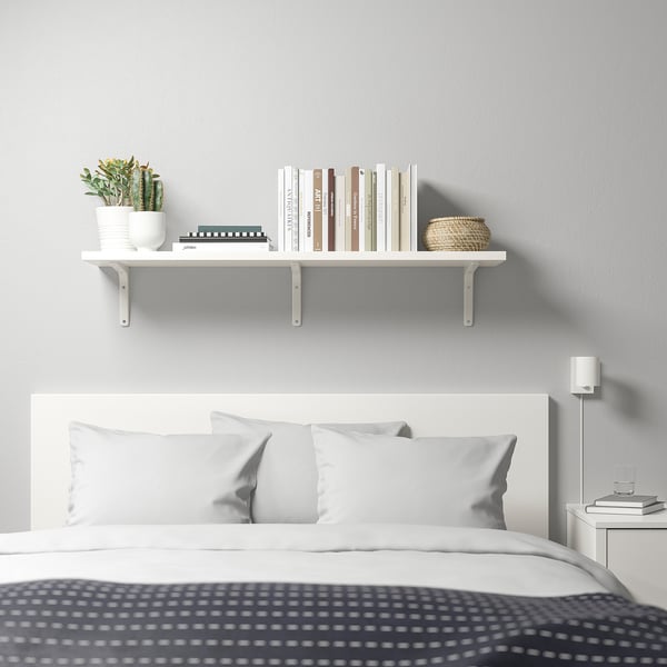 Une chambre minimaliste avec une étagère TOMTHULT au-dessus du lit, contenant des livres, une plante et un panier. Une table de chevet avec des livres et une lampe.