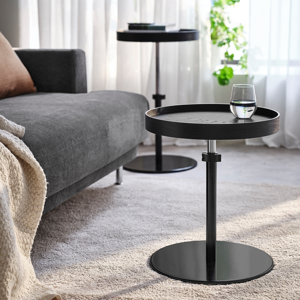 Table d’appoint à côté du canapé gris, réglable, avec un verre d’eau sur un support métallique noir.