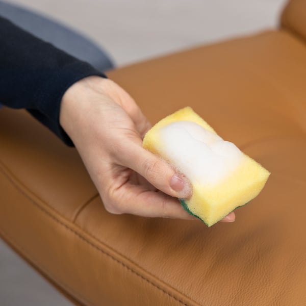 Une main tient une éponge jaune nettoyant une surface en cuir marron, démontrant la capacité des nettoyants pour cuir TRIXIG à nettoyer et à entretenir les meubles en cuir.