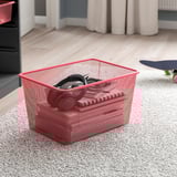 Le panier de rangement en maille rouge peut contenir de petits objets, idéal pour l’organisation. Positionné sur un sol recouvert de moquette avec une planche à roulettes à proximité.