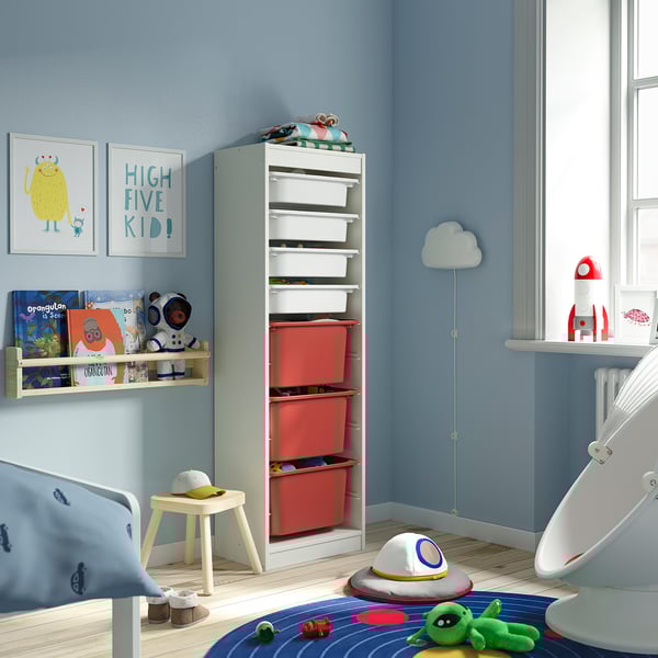 Chambre d’enfant avec unité de rangement TROFAST bleu clair en panneaux de particules et en plastique. Il est rempli de boîtes en maille rouge, plus petites qu’une grande fenêtre à proximité.