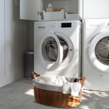 Une machine à laver blanche avec une porte ouverte, un panier à linge avec des draps à côté et des étagères de rangement au-dessus.