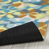 Gros plan d’un paillasson floral coloré avec un support en caoutchouc noir texturé et antidérapant sur un plancher en bois.