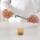 Personne pressant l’eau de la brosse à vaisselle. La brosse a un manche en bois et une presse en métal.