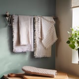 Des serviettes à rayures blanches avec des franges sont suspendues sur un support mural, mur bleu clair. Planche à découper en bois et gant de cuisine à proximité, plante verte à la fenêtre.