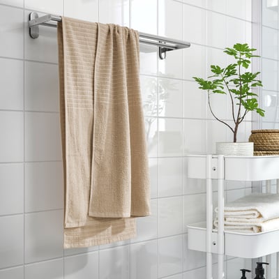 Une salle de bain avec une serviette VÅGSJÖN beige sur un support, des carreaux blancs et un support à trois niveaux avec des serviettes et une plante.