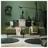 Salon vert avec canapé vert, coussins colorés, papier peint cactus et lampadaire.