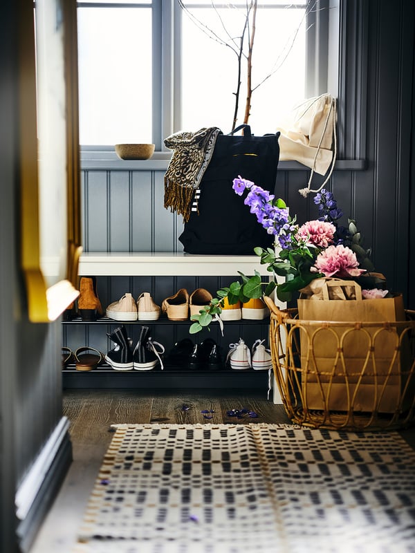 Entrée bien organisée avec sac à dos féminin noir, fleurs violettes, panier en osier et tapis à carreaux.