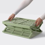 Personne tenant une boîte pliante VEDKLYV vert olive avec poignées, montrant comment la déplier sans effort.