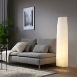 Un lampadaire VICKLEBY avec un abat-jour ovale en papier de riz, émettant une lumière douce à côté d’un canapé gris.