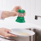 La main tient la brosse verte en libérant le savon sur une assiette sale. Évier en métal et robinet à proximité.