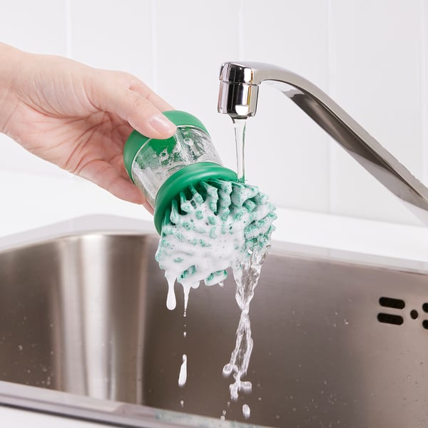 Une main tient une brosse verte qui dissipe le savon sous le robinet d’un évier.
