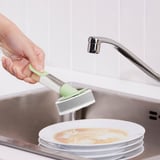 Une main tient une éponge avec une poignée remplie de savon au-dessus d’un évier. L’éponge presse le savon sur un plat sale en dessous.