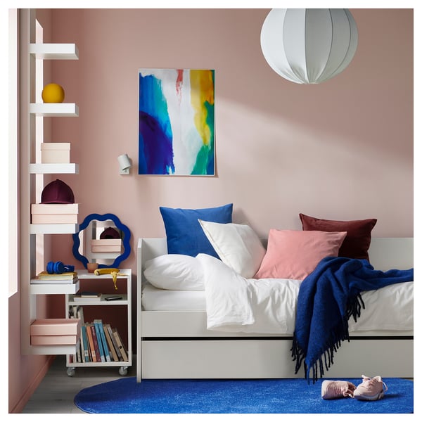 Chambre confortable avec lit de repos VIHALS blanc, oreillers colorés, tapis bleu, étagère avec des livres et de l’art abstrait sur mur rose.