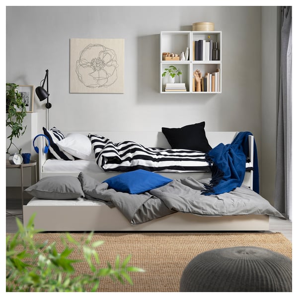 Chambre confortable avec un couvre-lit rayé blanc et gris, des accents bleus et une étagère murale. Des plantes et des œuvres d’art décorent l’espace.