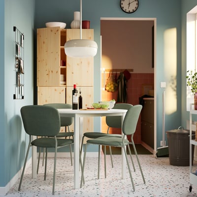 Salle à manger moderne avec table ronde VISALS blanche et chaises vertes, murs bleu clair, rangements en bois et sol moucheté.