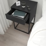 Table de chevet noire avec tiroir ouvert montrant des livres, à côté d’une horloge blanche et d’une vitre sur le dessus, à côté d’un lit blanc.