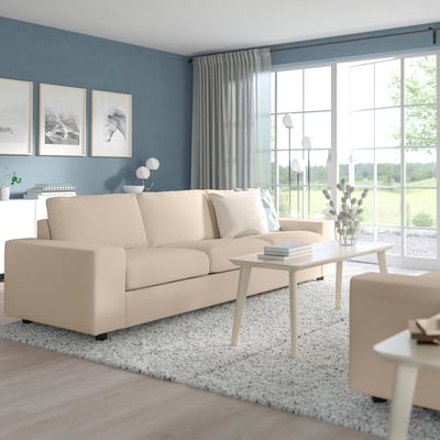 Salon moderne avec canapé beige VIMLE, table basse en bois clair, tapis blanc, mur bleu, grandes fenêtres, rideaux gris et lumière naturelle.