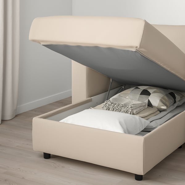 Canapé-lit beige ouvert, montrant un matelas épais et un compartiment de rangement avec couvertures et oreillers.