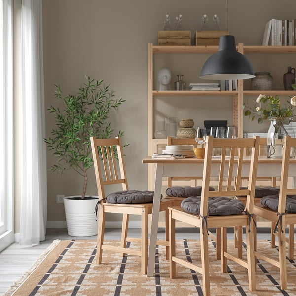 Salle à manger moderne avec chaises en bois et coussins gris, tapis lumineux, plante en pot et étagère avec décor. Les chaises ont des coussins en tissu recyclé gris foncé.