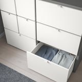 Commode blanche avec tiroirs lisses et pieds réglables. Un tiroir s’est ouvert, montrant les vêtements.