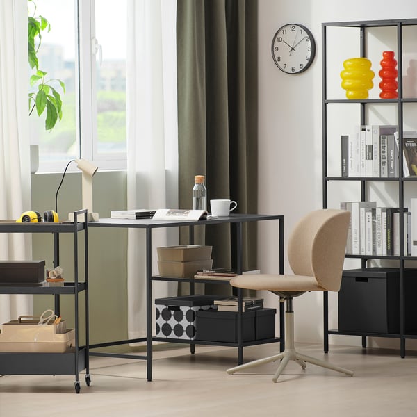 Bureau noir avec étagères en verre, lampe et chaise, près de la fenêtre, à côté d’une étagère colorée.