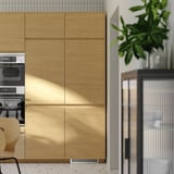 Armoires de cuisine modernes en bois avec poignées intégrées, design élégant en chêne clair, avec fours intégrés et plante en pot.