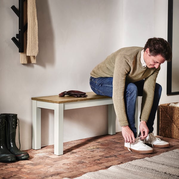 Person tying white trainers, bending near a wooden bench with black boots and gloves; a towel hangs on a hook nearby.