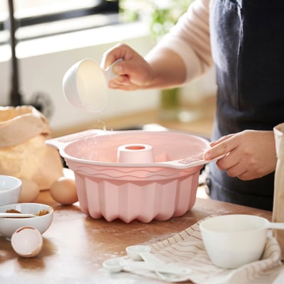 Baker preparing cake tin with flour, eggs, sugar nearby.
