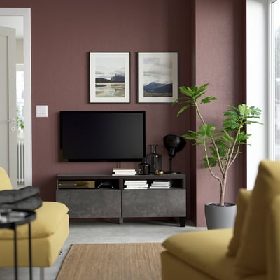 Modern living room with maroon walls, yellow sofa, flat-screen tv on BESTÅ stand with drawers, books, and tall plant. Two pictures above tv.