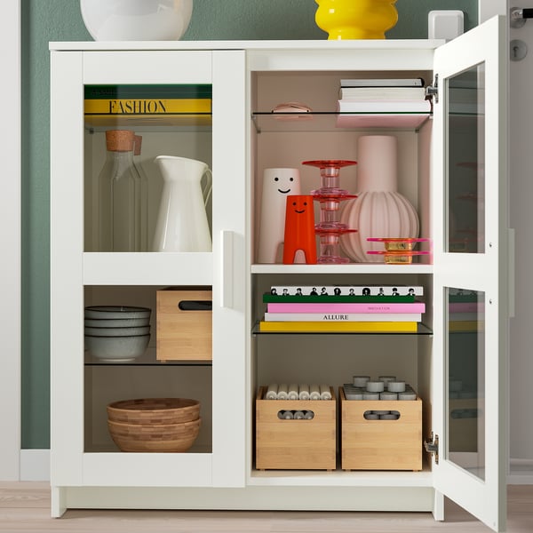 White BRIMNES cabinet with glass doors, storing vases, bowls, books, and boxes. Shelves are adjustable, showcasing items neatly.