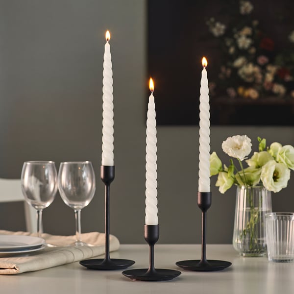 Three lit spiral candles in black holders on a table with glasses and flowers.