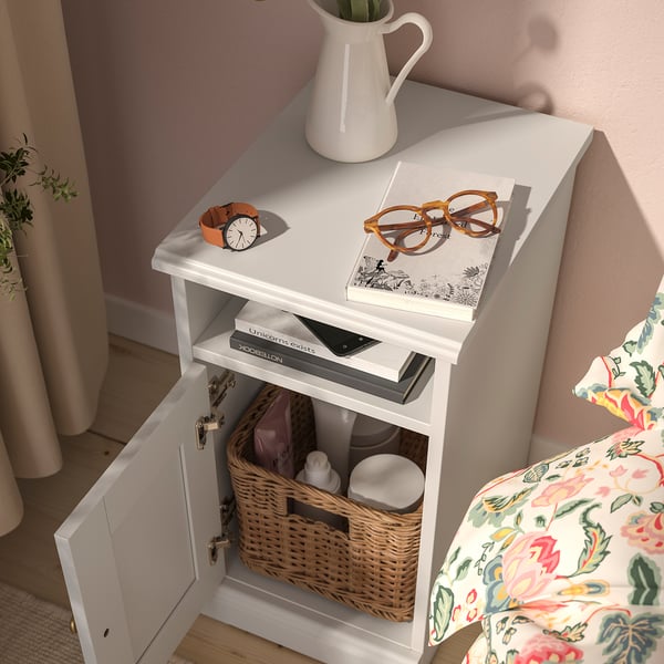White bedside table with open door revealing a wicker basket holding bottles. Top has books, glasses, and a clock.