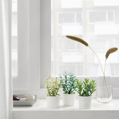 Windowsill with three potted artificial plants and a tall vase with dried grasses, bringing indoor nature without maintenance.