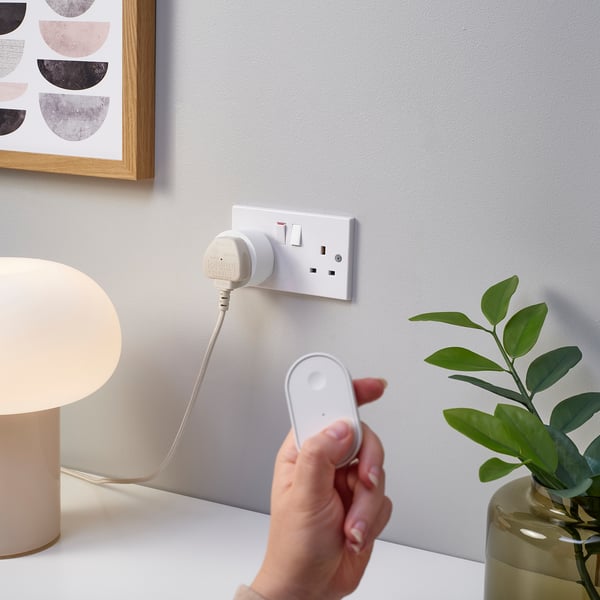 Hand holds remote aiming at smart plug. Plug is in wall socket, cable to lamp. Plant beside.