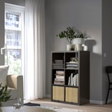 KALLAX bookshelf in grey by window, filled with books and a vase of flowers.