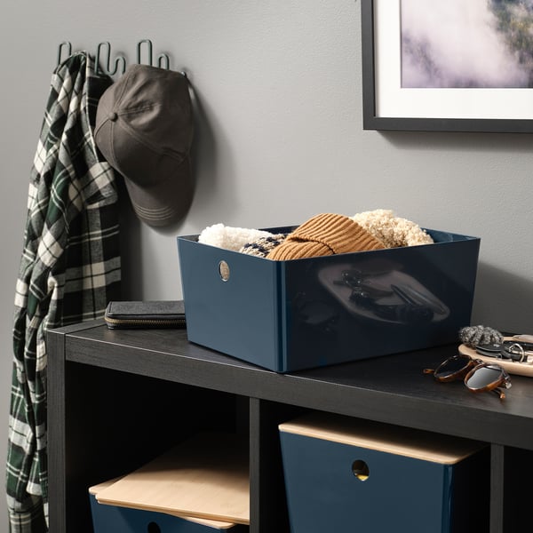 Dark blue storage box with wooden lid filled with clothes on dark shelf next to sunglasses and scarf.