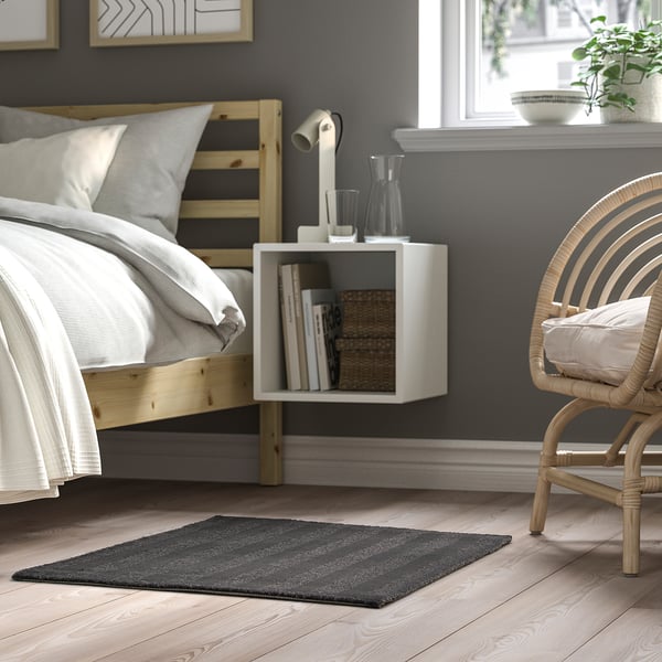 Modern bedroom with grey rug, wooden bed, white bedside table, wicker chair, plant, & wall art. Rug: dark grey with subtle stripes.