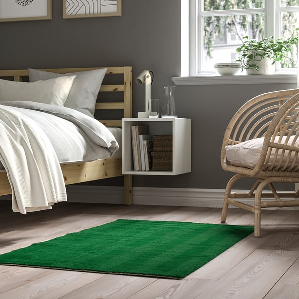 Cosy bedroom with LANDSVÄG green rug, soft low pile, stripe design. Rug beside bed, smaller than window, wicker chair atop.