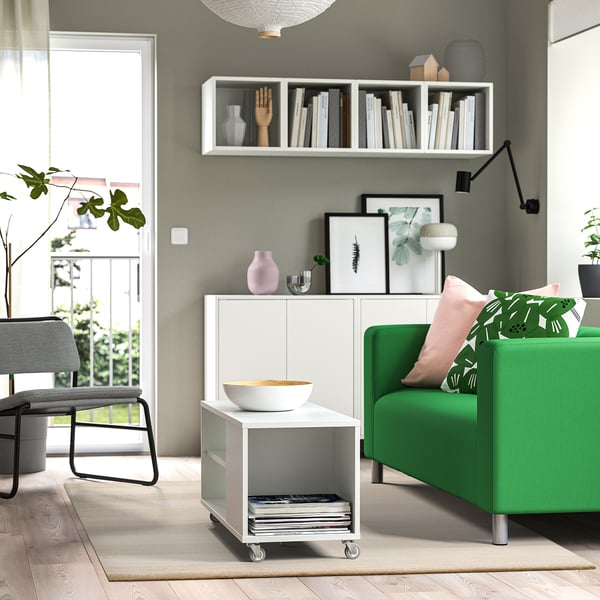 A modern living room a green couch with leaf-pattern pillows, a white low-pile rug, and a white LANDSVÄG storage unit with a bowl atop.